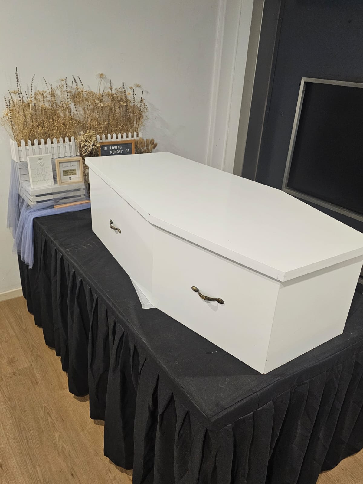 White custom-made pet casket resting on a draped table beside memorial flowers